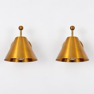 May include: A pair of wall-mounted light fittings featuring conical brass shades with a brushed finish. Each lamp has a central vertical stem topped with a small spherical finial, extending from a circular wall plate, with a light bulb visible inside.