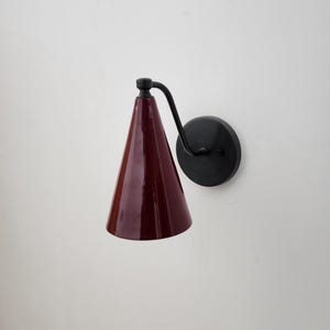 May include: A wall-mounted sconce with a conical, burgundy-colored shade. The shade is attached to a curved black arm and a circular black base. The sconce is designed for indoor lighting and has a modern aesthetic.