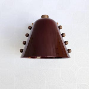 A Decorative Wine Colored Cone Wall Lamp with Handcrafted Brass Construction, Photographed to Highlight Its Compact Proportions, Tapered Shade, and Distinctive Rivet Details in A Mid Century Inspired Form, Frost Lights.