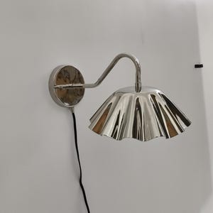 Könnte beinhalten: Silberfarbene Wandleuchte mit einem gerüschten, blütenblattartigen Schirm. Die Leuchte hat einen gebogenen Arm und eine kreisförmige Basis, und ein schwarzes Kabel verläuft die Wand hinunter. Die Leuchte ist an einer weißen Wand befestigt.