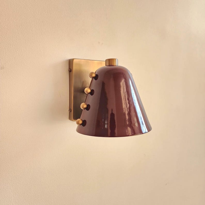 Handcrafted Wall Sconce with A Rich Wine Colored Tapered Cone Shade, Solid Brass Construction, and Decorative Rivet Details, Shown in A Compact Profile that Suits Mid Century Inspired Interior Lighting, Frost Lights.