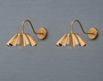 May include: A pair of wall-mounted light fixtures featuring a brushed brass finish. Each lamp has a curved arm extending from a circular wall plate to a wide, scalloped, petal-shaped shade that directs light downward over a single exposed light bulb.