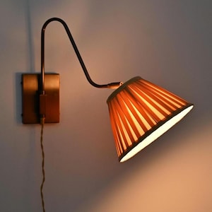 Op de afbeelding: Een wandlamp met een donkerbronskleurige metalen arm met een gebogen ontwerp en een rechthoekige basisplaat. De lamp heeft een geplooide, kegelvormige stoffen kap in een warme amberkleur, die een zachte gloed van de lichtbron uitstraalt.