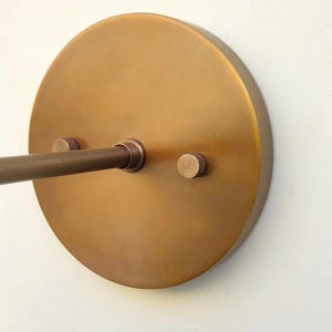 Könnte beinhalten: Nahaufnahme einer messingfarbenen Wandleuchte. Die Leuchte hat eine kreisförmige Rückplatte mit einer horizontalen Stange und zwei kleinen, runden Schrauben. Die Oberfläche hat einen warmen, metallischen Farbton.