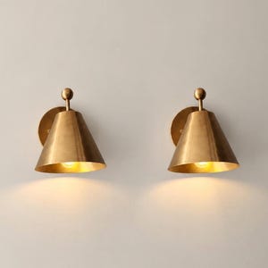 May include: A pair of wall-mounted sconces featuring a conical brass shade and a small spherical finial at the top. Each fixture is attached to a circular wall plate and emits a warm, downward-facing light from an internal bulb.