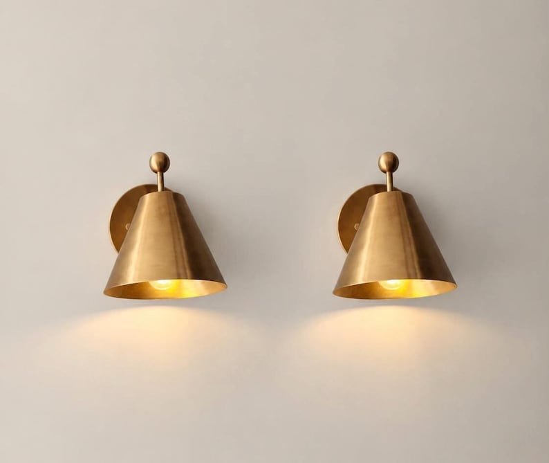 May include: A pair of wall-mounted sconces featuring a conical brass shade and a small spherical finial at the top. Each fixture is attached to a circular wall plate and emits a warm, downward-facing light from an internal bulb.