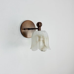May include: A wall-mounted light fitting with a bronze finish. It features a circular base, a curved arm, and a tulip-shaped shade made of translucent, off-white material with subtle veining. The fixture is designed to be mounted on a wall.