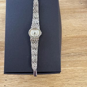 Puede incluir: Un reloj de pulsera plateado con una esfera blanca y una pulsera engastada con diamantes. El reloj está en una caja negra.