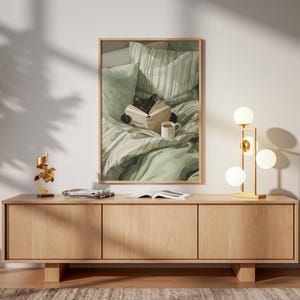 May include: A framed print depicts a black cat reading a book in bed with a cup of coffee. A wooden cabinet with three doors holds a gold lamp with spherical bulbs. A gold sculpture and open book sit on the cabinet.