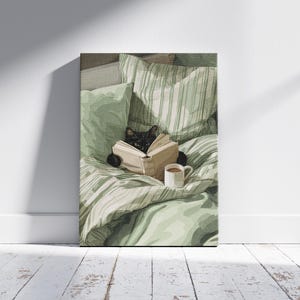 May include: A canvas print depicts a black cat reading a book in bed, surrounded by green pillows and a striped blanket. A white mug with a brown beverage sits nearby. The artwork is displayed against a white wall and wooden floor.