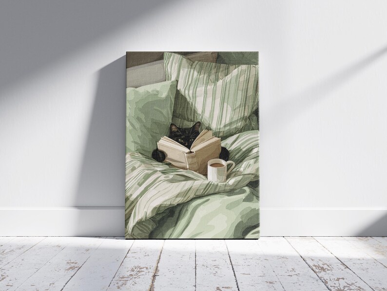 May include: A canvas print depicts a black cat reading a book in bed, surrounded by green pillows and a striped blanket. A white mug with a brown beverage sits nearby. The artwork is displayed against a white wall and wooden floor.