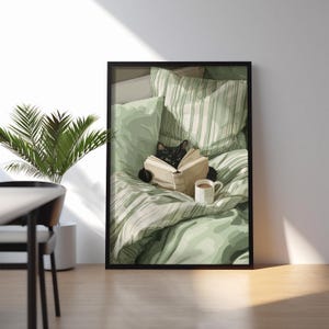 May include: A framed print depicts a black cat reading a book in bed. The cat is surrounded by green and white striped bedding and pillows. A white mug of coffee sits nearby. The artwork is displayed in a room with a plant.