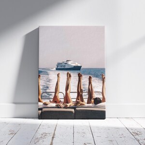May include: A canvas print depicts a sunny day on a boat. Several pairs of legs are raised in the air, with a ferry boat in the background on the blue water. The print is displayed against a white wall and a wooden floor.