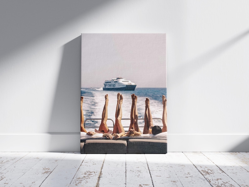 May include: A canvas print depicts a sunny day on a boat. Several pairs of legs are raised in the air, with a ferry boat in the background on the blue water. The print is displayed against a white wall and a wooden floor.