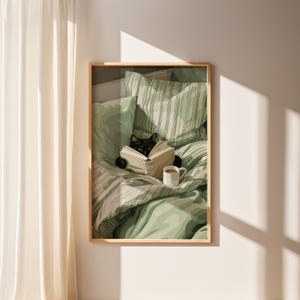 May include: A framed print depicts a black cat reading a book in bed. The cat is nestled among green and white striped bedding and pillows. A white mug with a brown beverage sits nearby. The artwork is in a light wooden frame.