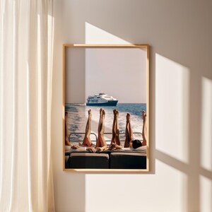 May include: A framed print featuring a boat scene with several pairs of legs raised in the air. A large ferry boat is visible in the background, sailing on the blue ocean. The image is framed in a light wood frame.