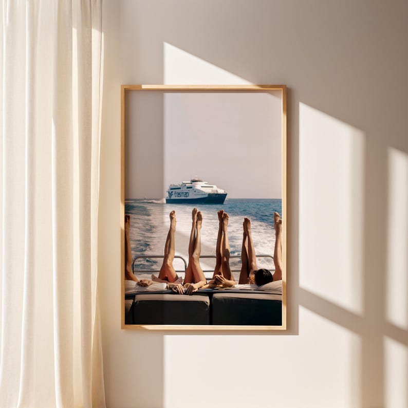 May include: A framed print featuring a boat scene with several pairs of legs raised in the air. A large ferry boat is visible in the background, sailing on the blue ocean. The image is framed in a light wood frame.