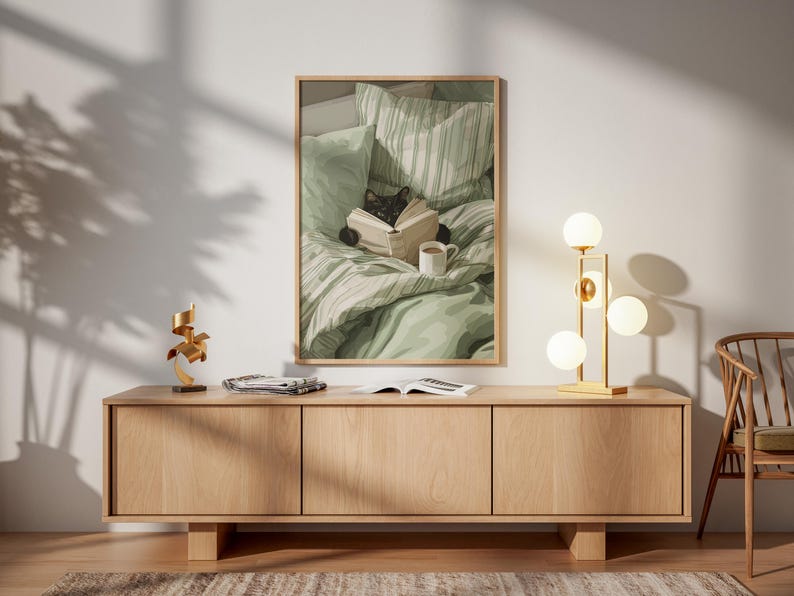 May include: A framed print depicts a black cat reading a book in bed with a cup of coffee. A wooden cabinet with three doors holds a gold lamp with spherical lights. A gold sculpture and open book are on the cabinet.