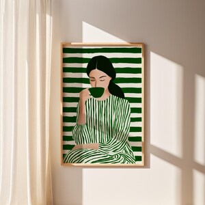 May include: A framed art print featuring a woman sipping from a green cup. The woman is depicted in a green and white striped dress, set against a green and white striped background. The artwork has a minimalist, modern aesthetic.