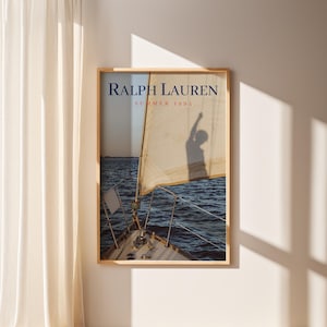 May include: A framed print featuring a sailboat on the water with a person's shadow on the sail. The print has the text "RALPH LAUREN SUMMER 1994" in navy blue and red. The frame is a light wood color.