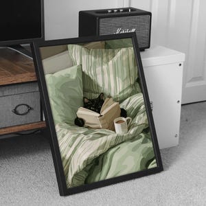 May include: A framed print depicts a black cat nestled in a bed with green striped bedding, engrossed in a book. A white mug sits nearby. The artwork is in a black frame, suggesting a cozy, relaxing theme.