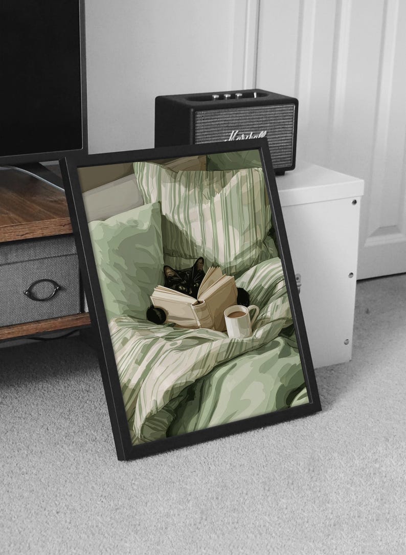 May include: A framed print depicts a black cat nestled in a bed with green striped bedding, engrossed in a book. A white mug sits nearby. The artwork is in a black frame, suggesting a cozy, relaxing theme.