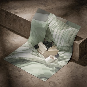 May include: A black cat is nestled among pillows and a blanket, engrossed in a book. A white mug with a dark beverage sits nearby. The bedding features a soft green color with striped and solid patterns, creating a cozy scene.