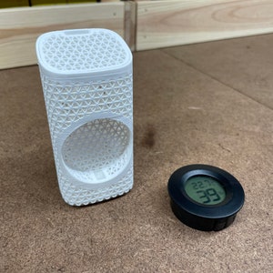 May include: A white 3D printed cylindrical object with a lattice pattern and a round opening. A black round digital hygrometer with a display showing 22.99 degrees.