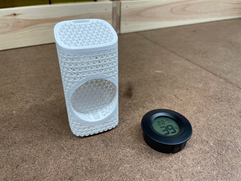 May include: A white 3D printed cylindrical object with a lattice pattern and a round opening. A black round digital hygrometer with a display showing 22.99 degrees.