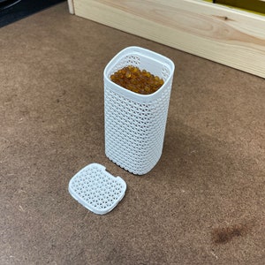 May include: A white plastic container with a lid, filled with small, round, amber-colored beads. The container has a lattice-like design.