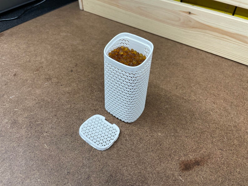 May include: A white plastic container with a lid, filled with small, round, amber-colored beads. The container has a lattice-like design.