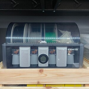 May include: A black plastic box with a clear lid containing six spools of filament for a 3D printer. The spools are labeled "AMS" and have a digital display showing the temperature.