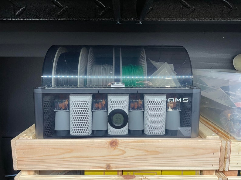 May include: A black plastic box with a clear lid containing six spools of filament for a 3D printer. The spools are labeled "AMS" and have a digital display showing the temperature.