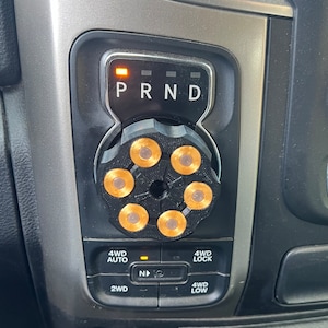 May include: A black gear shift knob with six gold bullet casings arranged in a circular pattern. The knob is installed in a vehicle's dashboard, near the gear shift indicator with the letters P, R, N, and D.