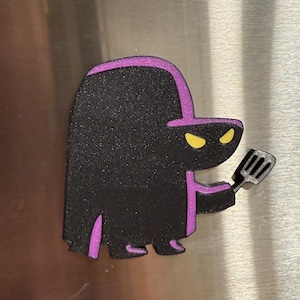 May include: A black and purple cartoon character magnet with yellow eyes, holding a spatula. The character has a long, rounded body and a hat. The magnet is likely for a refrigerator or other metal surface.