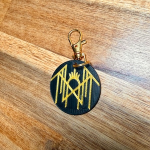 May include: A black and gold keychain with a circular pendant. The pendant features a gold geometric design and a gold clasp. The keychain is on a wooden surface.
