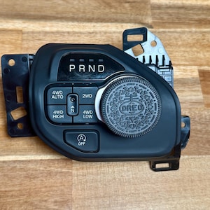 Pode incluir: Uma consola de mudança de marchas de carro preta com um mostrador prateado em forma de Oreo. A consola tem botões marcados "PRND", "4WD AUTO", "2WD", "4WD HIGH", "4WD LOW" e um botão "A OFF".