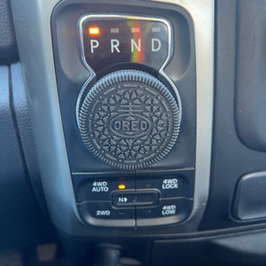 Könnte beinhalten: Nahaufnahme der Gangschaltung eines Autos. Die Gangwahlanzeige zeigt "PRND" mit einem orangefarbenen Licht. Ein Oreo-Keks ist auf dem Schalthebelknopf platziert. Die Konsole hat auch Tasten für 4WD AUTO, 4WD LOCK, 2WD und 4WD LOW.