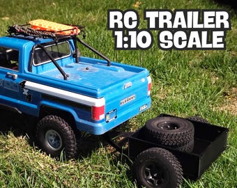 May include: A blue and white pickup truck with a black roll cage and a roof rack is towing a black trailer with a spare tyre on the bed. The truck has the word "Elegant" on the back. The text "RC TRAILER 1:10 SCALE" is in the upper right corner of the image.