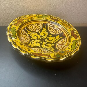 May include: A round ceramic plate with a scalloped edge, featuring a vibrant yellow base color. The center displays a dark brown and yellow floral-inspired motif, surrounded by geometric patterns and green and brown leaf-like accents.