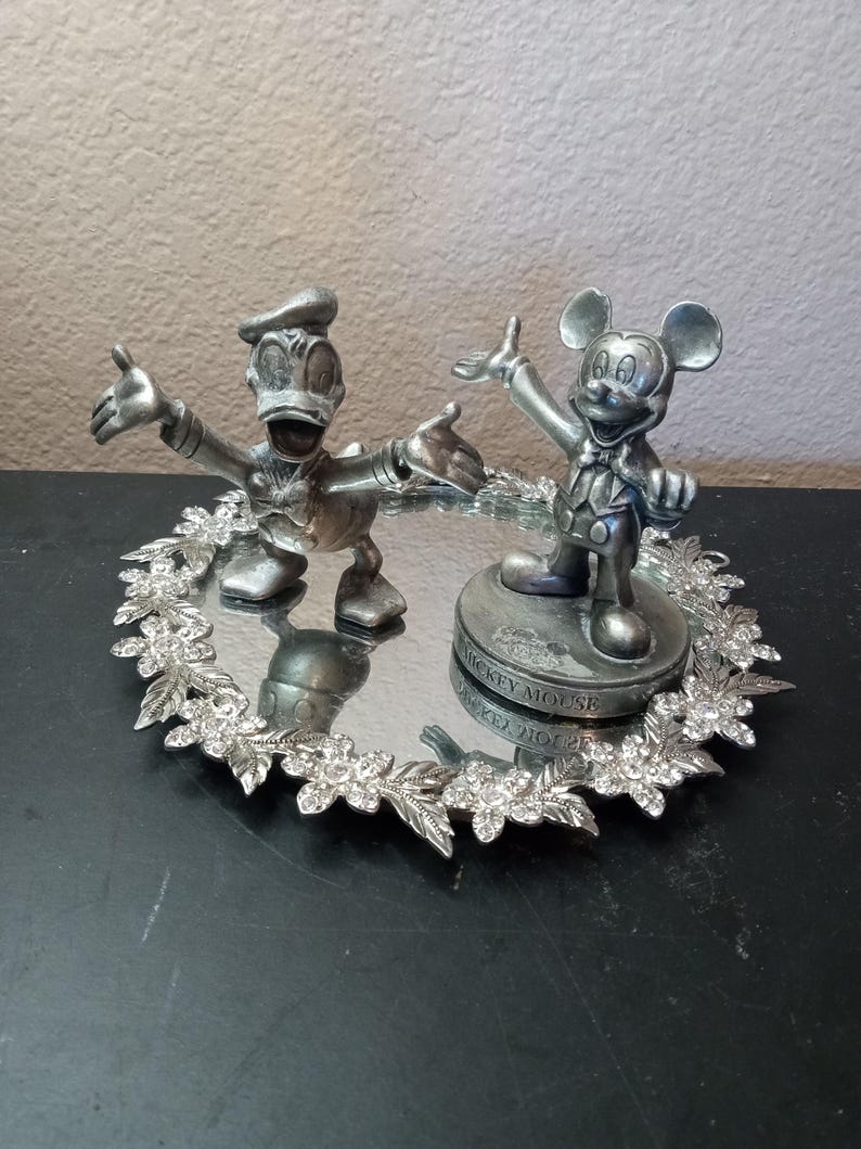 Puede incluir: Una bandeja decorativa plateada con figuras de Pato Donald y Mickey Mouse. Las figuras est&aacute;n posando con los brazos extendidos, sobre una superficie espejada rodeada por un borde floral adornado con pedrer&iacute;a. La base de Mickey Mouse dice "Mickey Mouse".