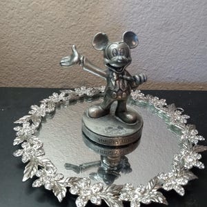 Puede incluir: Figura plateada de Mickey Mouse sobre una bandeja espejada. La bandeja est&aacute; rodeada por una guirnalda floral decorativa con detalles de cristal transparente. La figura est&aacute; sobre una base con texto.
