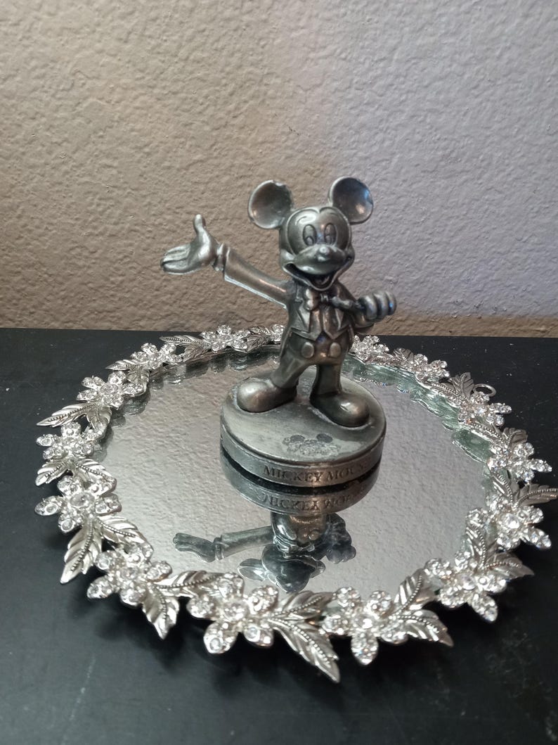 Puede incluir: Figura plateada de Mickey Mouse sobre una bandeja espejada. La bandeja est&aacute; rodeada por una guirnalda floral decorativa con detalles de cristal transparente. La figura est&aacute; sobre una base con texto.
