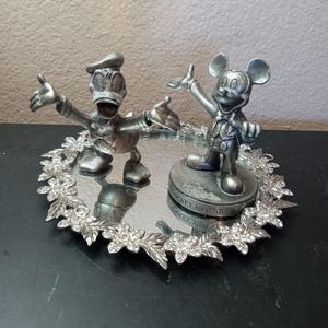 Puede incluir: Una bandeja decorativa plateada con figuras de Mickey Mouse y el Pato Donald. La bandeja tiene una superficie de espejo y est&aacute; rodeada por un dise&ntilde;o floral con detalles de cristal. La figura de Mickey Mouse est&aacute; sobre una base que dice "Mickey Mouse".