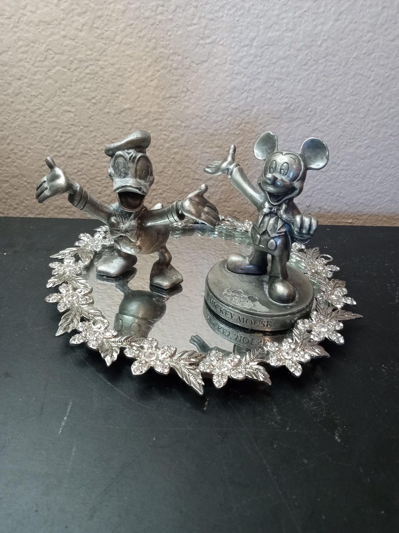Puede incluir: Una bandeja decorativa plateada con figuras de Mickey Mouse y el Pato Donald. La bandeja tiene una superficie de espejo y est&aacute; rodeada por un dise&ntilde;o floral con detalles de cristal. La figura de Mickey Mouse est&aacute; sobre una base que dice "Mickey Mouse".