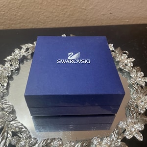 Könnte beinhalten: Eine quadratische, dunkelblaue Swarovski-Schmuckschatulle mit dem weißen Schwan-Logo und dem Markennamen auf dem Deckel. Die Schatulle steht mittig auf einem runden, silberfarbenen Dekotablett mit einem detaillierten Blumen- und Blattmuster.