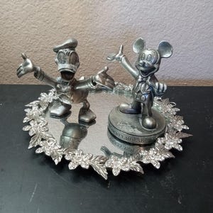 Puede incluir: Un juego de figuras plateadas de Mickey Mouse y el Pato Donald. Mickey Mouse est&aacute; sobre una base con texto, y el Pato Donald est&aacute; a su lado. Est&aacute;n sobre una bandeja espejada con un borde floral y cristalino.