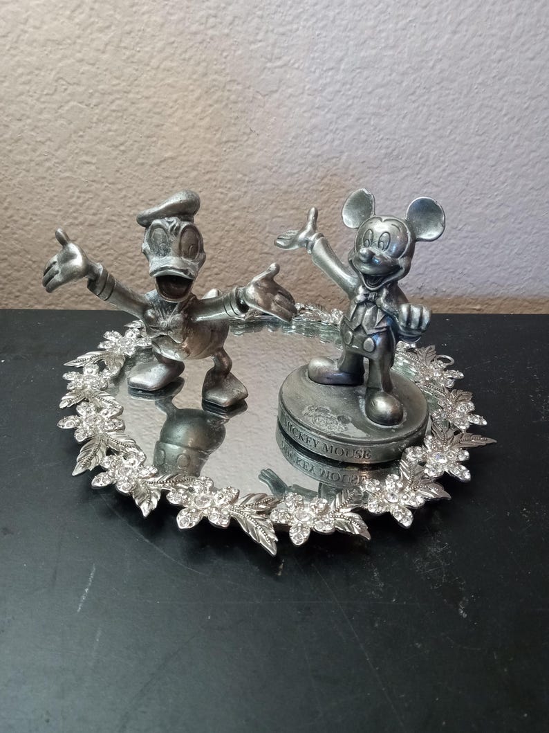 Puede incluir: Un juego de figuras plateadas de Mickey Mouse y el Pato Donald. Mickey Mouse est&aacute; sobre una base con texto, y el Pato Donald est&aacute; a su lado. Est&aacute;n sobre una bandeja espejada con un borde floral y cristalino.