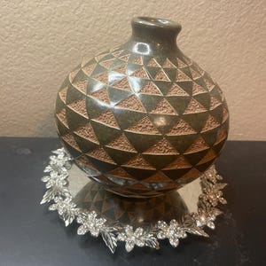 May include: A round, dark green ceramic vase with a textured geometric pattern of alternating smooth and rough triangles. The vase sits on a circular silver-toned metal base decorated with a repeating floral design featuring clear embedded crystals.