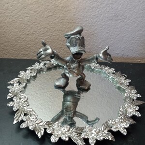 Puede incluir: Una figura plateada de Donald Duck con los brazos extendidos se encuentra sobre una bandeja ovalada con espejo. La bandeja est&aacute; bordeada con un dise&ntilde;o floral adornado con cristales transparentes. El reflejo de la figura es visible en el espejo.